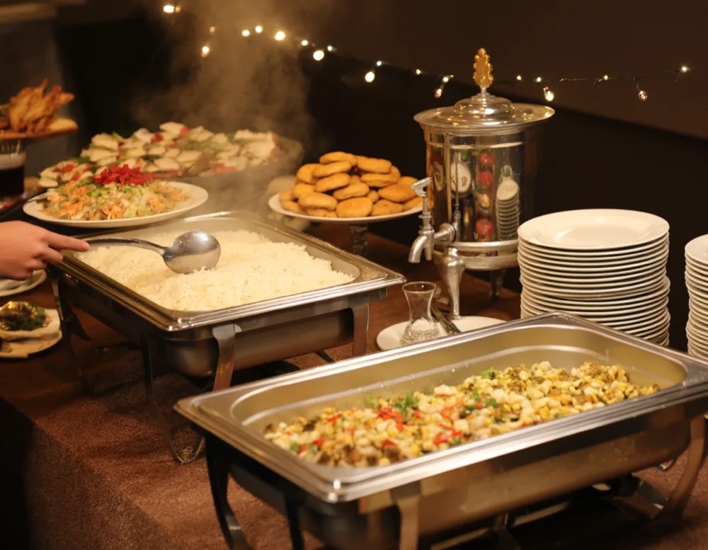 A self-service buffet table at a community-hall celebration with trays of rice, salads and pastries, a stack of plates and a tea urn