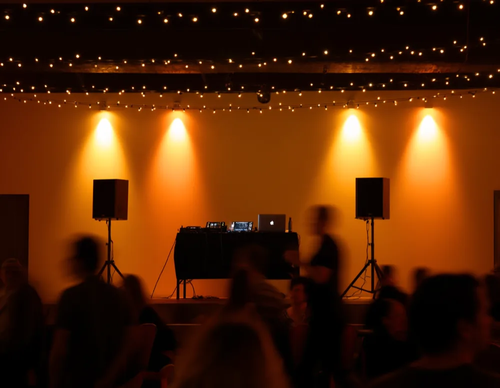 A small DJ booth with two speakers at the end of a community hall, warm uplighters on the wall