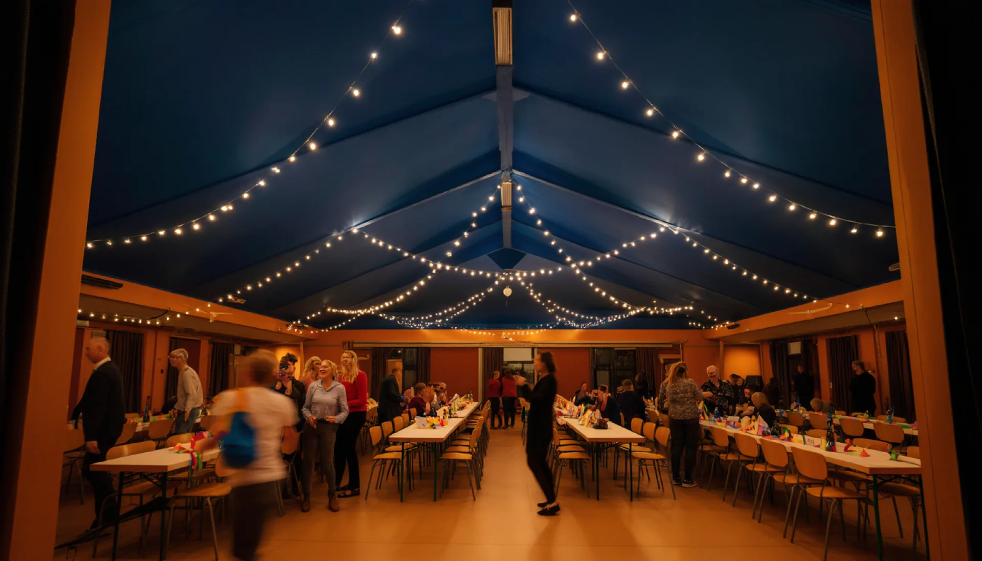 Guests laughing between long laid tables under string lights in a community hall in Herne