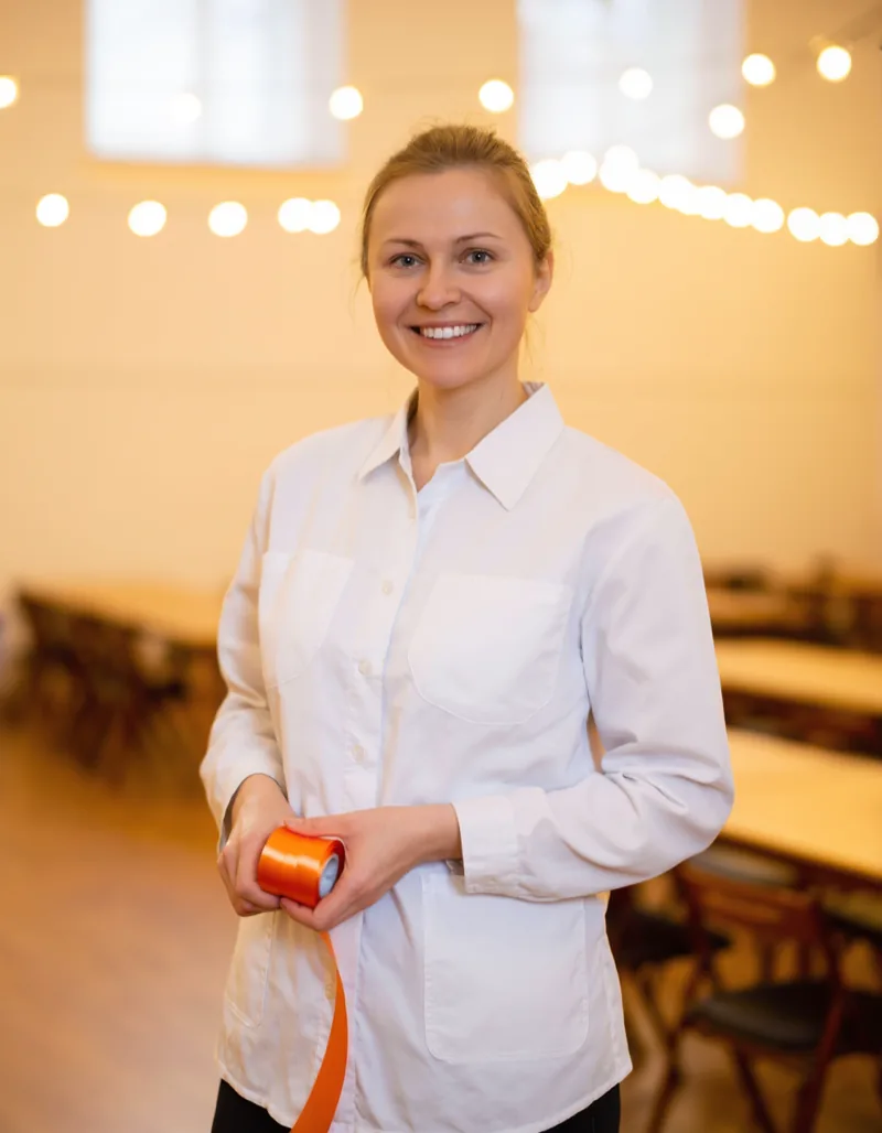 A member of the Paketfest set-up crew holding a roll of orange ribbon in a hall being decorated, string lights behind her